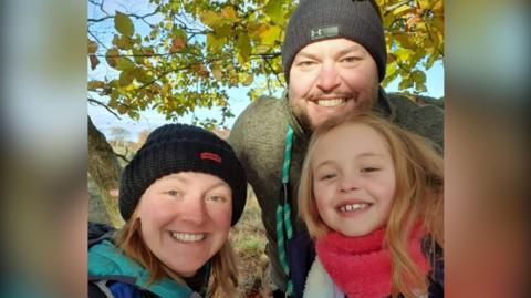 Library campaigner Kirsty Edwards wearing black hat and blue hooded jacket smiling in a selfie with her daughter and husband. Daughter is also smiling and wearing a pink coloured scarf. Kirsty's husband is also smiling and wearing a black hat. He has a beard and appears to have a dog lead over his shoulder. 