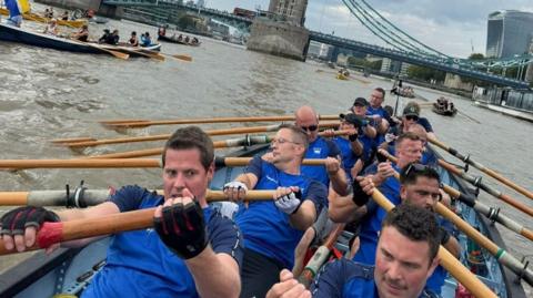 Men in blue t shirts row with intense expressions on their faces, with Tower Bridge in the background