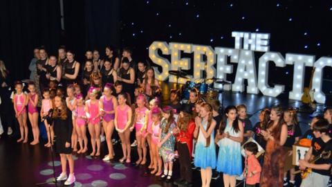 A group of young girls wearing colouful dresses on a stage. Behind them is an ulluminated sign which reads 'The Seb Factor'