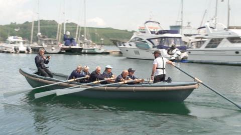 Six rowers row a grey boat in a harbour