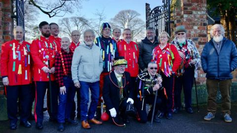 The full group of Redcar Sword Dancers with many in red uniforms with badges and ribbons attached. Two men in dark uniforms and white gloves are kneeling down at the front.