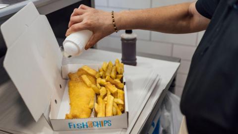 Over the shoulder shot of a worker holding a salt pot over a portion of fish and chips. The food is inside a carboard box with a lid.
