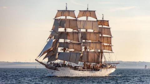 Tall cream-coloured ship - Sorlandet - with masts and beige sails at sea, with land and wind turbines in the distance.