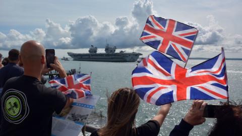 People waving union flags on the shore with HMS Prince of Wales warship sailing away in the distance