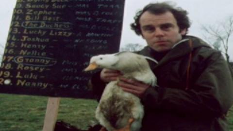 Man holding a duck while standing in front of a bookie's board in Caldecott.