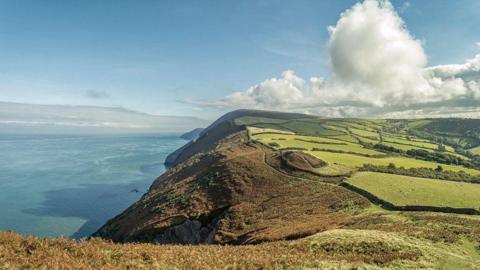 Looking east from Little Hangman to Great Hangman on a fine day, Girt Down, Combe Martin, Exmoor National Park. A sunny day with various vegetation on the coastal section. Blue sea and blue sky with a few white clouds.