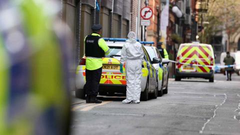 A police officer and a forensic investigator stand close to two police cars which are inside a cordon