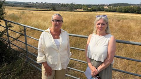 Two people stand by a farm gate overlooking a field of tall, dry grass, with rolling countryside and farmland in the background.