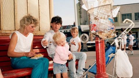 A 1980s photo of a family eating fish and chips, with piles of paper rubbish in the foreground