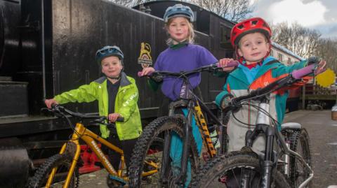 Three children wearing outdoor clothes and bicycle helmets stand in front of three bikes. In the background, a steam train carriage can be seen.