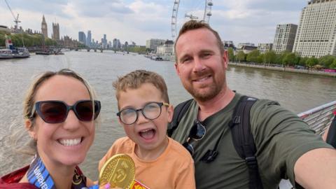 a mum and dad and child in London with the River Thames and the london Eye in the background. The mum, with blonde hair and sunglasses, is holding a London Marathon medal. Her son is wearing glasses and an orange t-shirt and her partner has a beard and is wearing a green t-shirt and black backpack