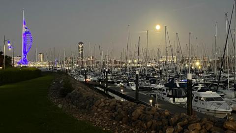 Multiple boats can be seen in Portsmouth Harbour under the moonlight, with Portsmouth's skyline illuminated in the background 