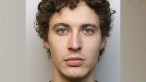 A mugshot of a man with short, curly brown hair. He is staring blankly at the camera in front of a grey background.