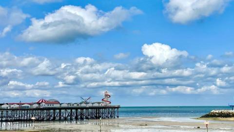 A pier with a helter skelter on the end goes out into the sea with blue sky and big white clouds behind