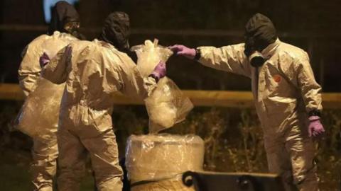 Officers wearing white full-body PPE inspecting bins. They are wearing purple gloves.