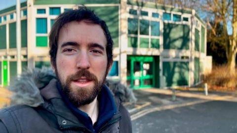 Joe Peall - who has dark hair and a beard - stood in front of the Polygon Centre, which is green and shaped like a decagon.