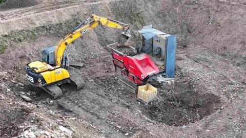 A yellow digger scoops waste into a red machine, where it is crushed and tipped into a hole.