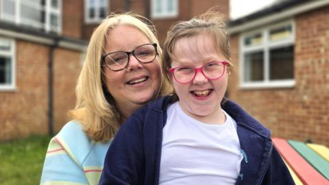 Claire and Katie, sitting outside and smiling at the camera. On the left Claire is wearing black rimmed glasses and and a light blue and green jumper. Sitting on her lap is Katie, who has pink rimmed glasses, whilst wearing a dark blue fleece over a light violet t-shirt.
