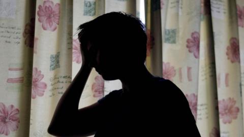 A silhouette of a woman holding her hand against her head. She is standing in front of floral curtains which are drawn. 