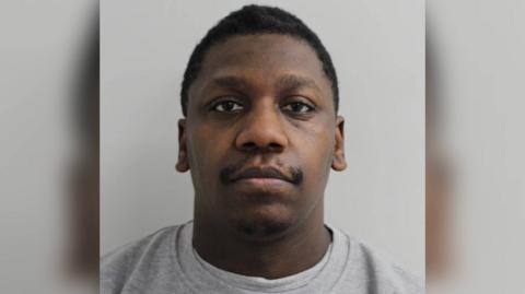 Jazz Reid, in a police custody shot, looking straight at the camera, wearing a light grey top