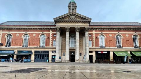 The front of Peterborough Town Hall. It is a two-storey red brick building with shops on the ground floor on either side of a pillared portico which rises the full two storeys and up into the roof, above which is a cupula. There are tall arched windows on the first floor above the shops. In front is an expanse of pavement.