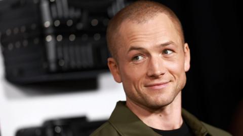 Taron Egerton attends the premiere of Netflix's  Carry-On at The Egyptian Theatre Hollywood on December 5, 2024. He is wearing a khaki jacket and black t-shirt. He has a shaved head and is smiling to the left of the camera. 