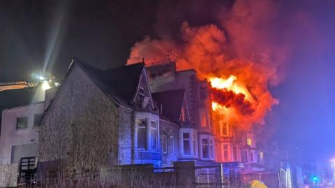 A building with huge orange flames pouring out the front. Thick smoke fills the night sky. The property on fire is attached to neighbouring buildings. A cherry picker is at the same height as the top floor of the building. A powerful torch on the cherry picker is shining a light into the night sky.
