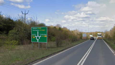 Street view of the A505 on a dry day. The straight flat stretch of road has two lanes with traffic flowing in opposite directions. There is a large green sign to the left pointing to various locations at the upcoming roundabout.
