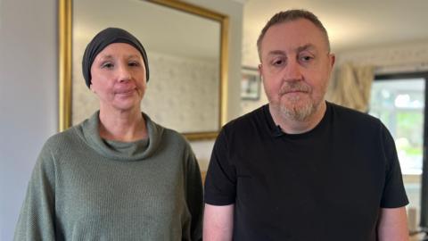 Caroline and Chris look into the camera with sad expressions. Caroline's head is wrapped in a blue scarf, and she wears a light green jumper. Chris has a pale moustache and is wearing a black t-shirt. 