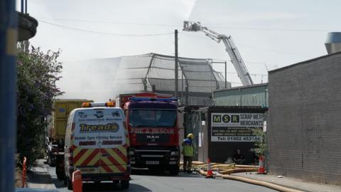A hydraulic platform can be seen in a recycling yard which is the scene of a large fire. Outside the building a West Midlands fire engine is parked on the road, also a van for a mobile tyre fitting service. One firefighter can be seen wearing a yellow helmet and high vis jacket.  