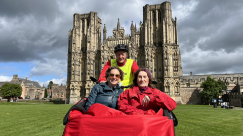 Two ladies tucked up under a blanket on a trishaw with a pilot behind in in vis clothing and helmet with a backdrop of Wells Cathedral