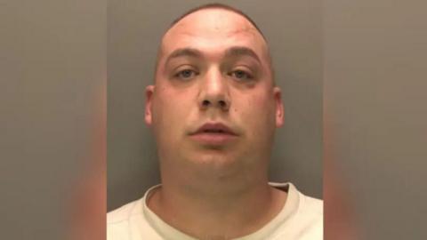 A police head shot of Joel who has a buzz cut hairstyle and is clean shaven. He is wearing a white t-shirt and is looking into the camera.
