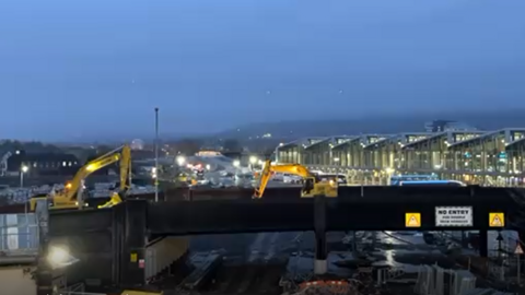 Two yellow diggers on top of the Boyne Bridge in Belfast city centre.  The new Grand Central Station is in the background 