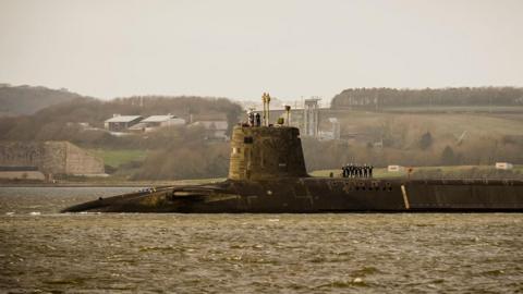 A submarine coming into port in Plymouth. There are officers standing in a line on the main body of it and a couple on the turret. It is a grey day and the sea looks brown.
