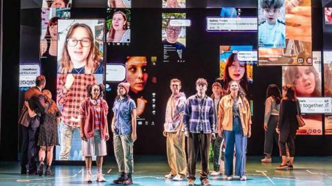 The cast of Dear Evan Hansen stand on the stage to sing You Will Be Found. Behind them are images of people appearing on screens on social media platforms. The six perfomers in the middle of the group are singing towards the audience. The other four characters are looking at the screens, two are standing upstage right and two upstage left.