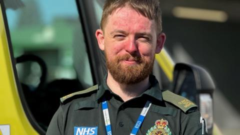 A paramedic wearing a dark green uniform which has an NHS label on. It also has an ambulance badge on it and he is also wearing a blue and white NHS lanyard. The man has auburn hair and and a beard and is smiling at the camera. 