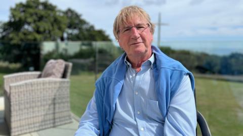 Chris Brock is sitting in a chair in on a sunny decking. He is wearing a blue shirt with a blue jumper draped round his shoulders. A glass panel separates the decking from the green grass below. An empty wicker chair is behind him.