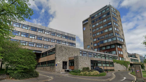 A grey/brown building at least 11 storeys high with windows. It is connected to a smaller two or three storey building of the same material on one side. On the other side connected to the smaller building is a taller building. A pathway leads up to the buildings.