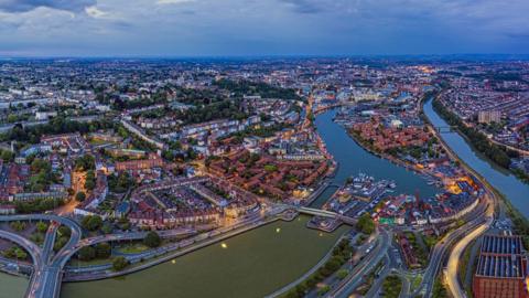 The image shows an aerial view of Bristol, with the Floating Harbour in the foreground and the Cumberland Basin also visible. The houses and green spaces of Clifton Wood are also visible, as is the River Avon and much of the centre. The image is taken at dusk and many points of light are visible