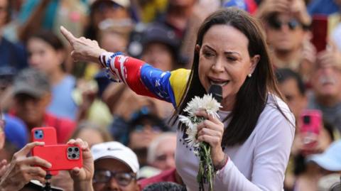Maria Corina Machado, a lady with brown hair and wearing a white t-shirt holds a microphone with flowers as a crowd stands behind her.