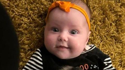 Dahlia-Rose Gartshore, a three-month-old baby girl looking at the camera, with a slight smile. She has an orange band on her head with a bow.