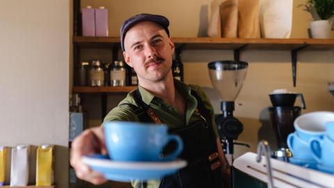 A barista extends a freshly brewed cup of coffee to the camera