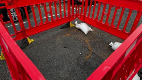 A picture of a tiny hole in tarmac in a road with red crash barriers around it, with two sandbags holding one of the four down.