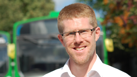 MP Tom Collins in front of a green bus. He has short sandy hair and is wearing glasses and a white shirt.