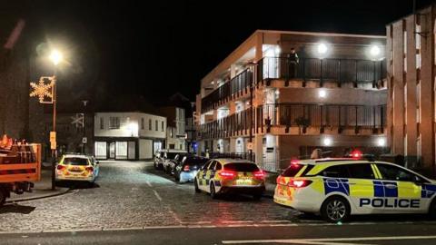 Three police vehicles parked near the scene. They are parked on a cobbled street. A block of flats with a black balcony is to the left. A police officer is stood outside on the second floor of the block of flats. It is dark. One street light is on. 