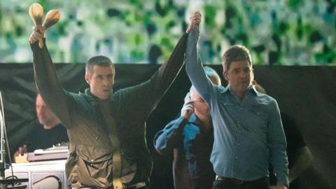 Two men raise each others hands. The man on the left is wearing a dark green raincoat and holds two shakers in his right hand. The man on the right wears a blue denim shirt, top button un-done, They both have short brown hair. Behind them are several men who stand in front of a screen displaying green patterns. 