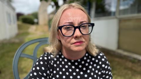 Julie Hogg looking into the camera while sitting outside hospital buildings. She has cropped blond hair, thick rimmed black glasses and is wearing a black top with white polka dots.