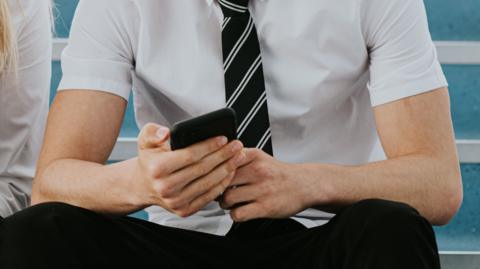 A male secondary school pupil sitting on a flight of stairs. He is holding a mobile phone and is showing it to a girl sitting next to him. He is wearing a short-sleeved white shirt and a black striped tie. Their faces are not visible.