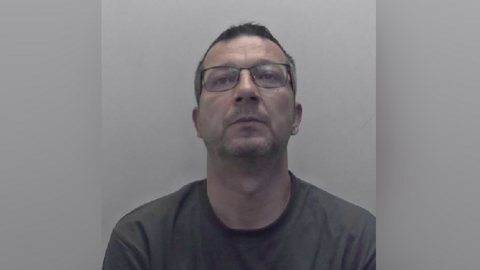 A mugshot of a man in a grey sweatshirt, with dark hair and glasses.