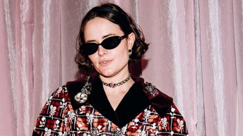 Kelley Heyer, a woman, at a fashion show, wearing black sunglasses and a red and black patterned outfit with a black v-neck and white flowers embroidered into the outfit. She is wearing a clasp around her neck, with a pink background behind her.